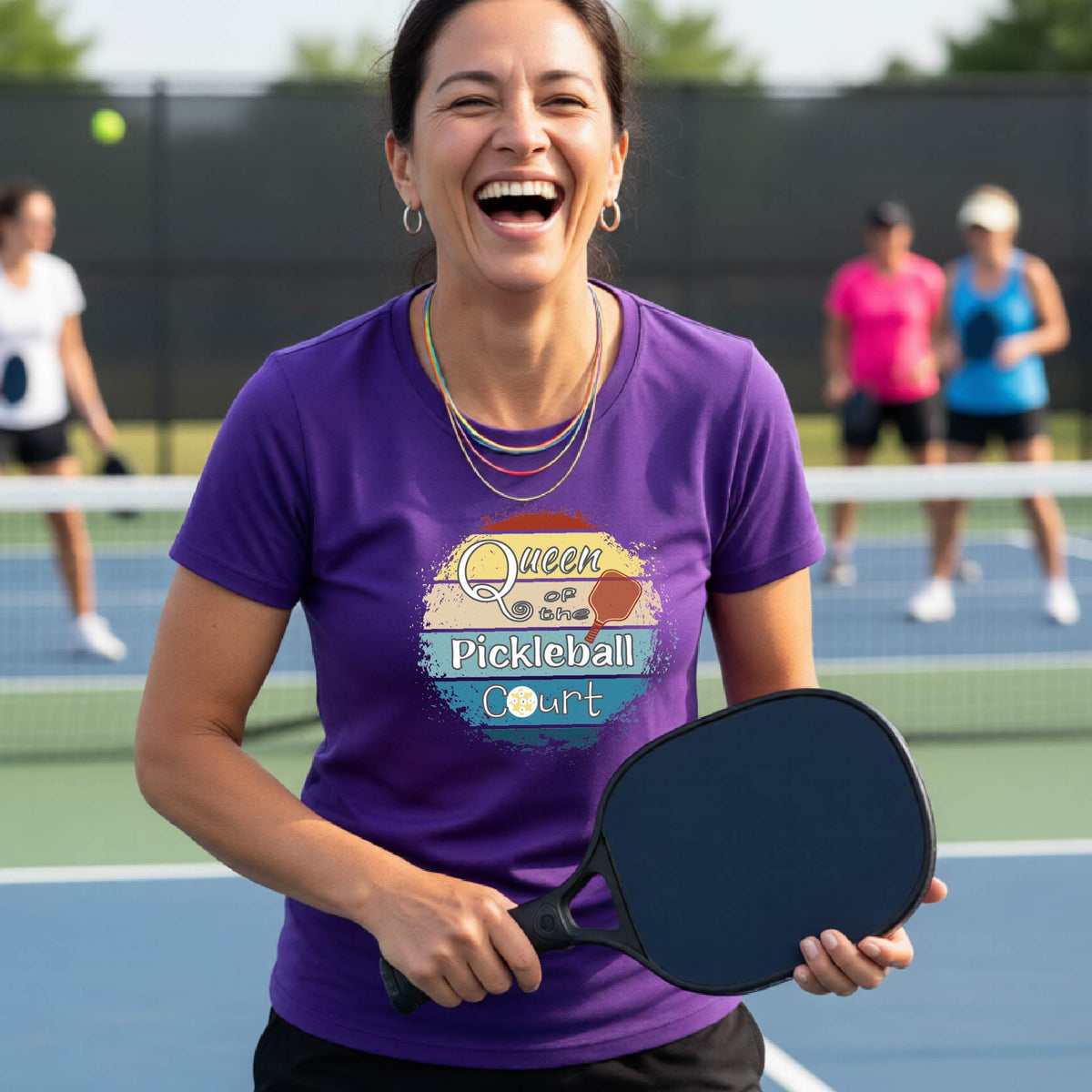 A woman on a pickleball court wearing a purple t-shirt with a 'Queen of the Pickleball Court' retro sunset graphic, holding a paddle and smiling. Other players are visible in the background on the court.