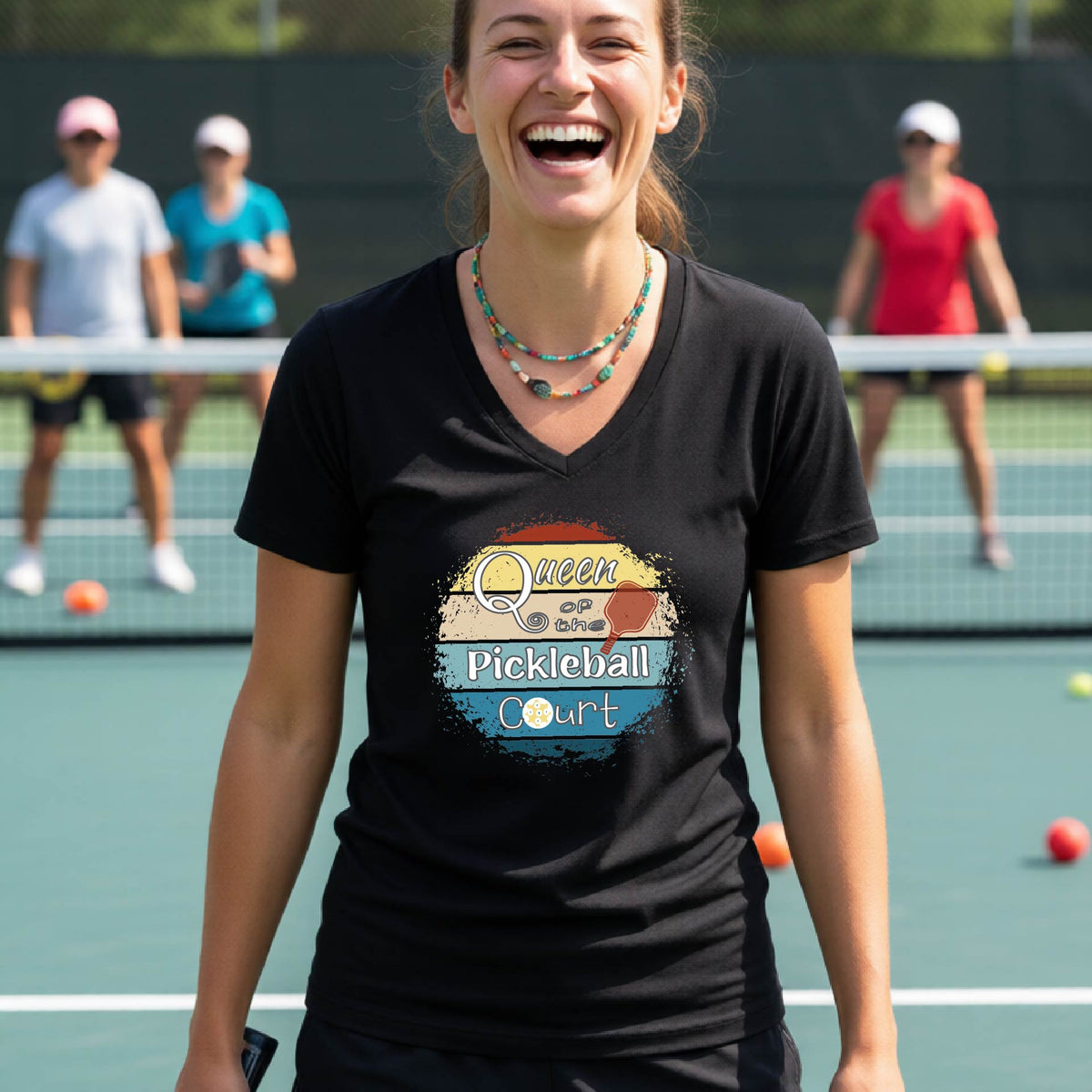 A woman on a pickleball court wearing a black V-neck t-shirt with a colorful retro-style graphic that reads 'Queen of the Pickleball Court' with a retro sunset illustration. Other players are blurred in the background.