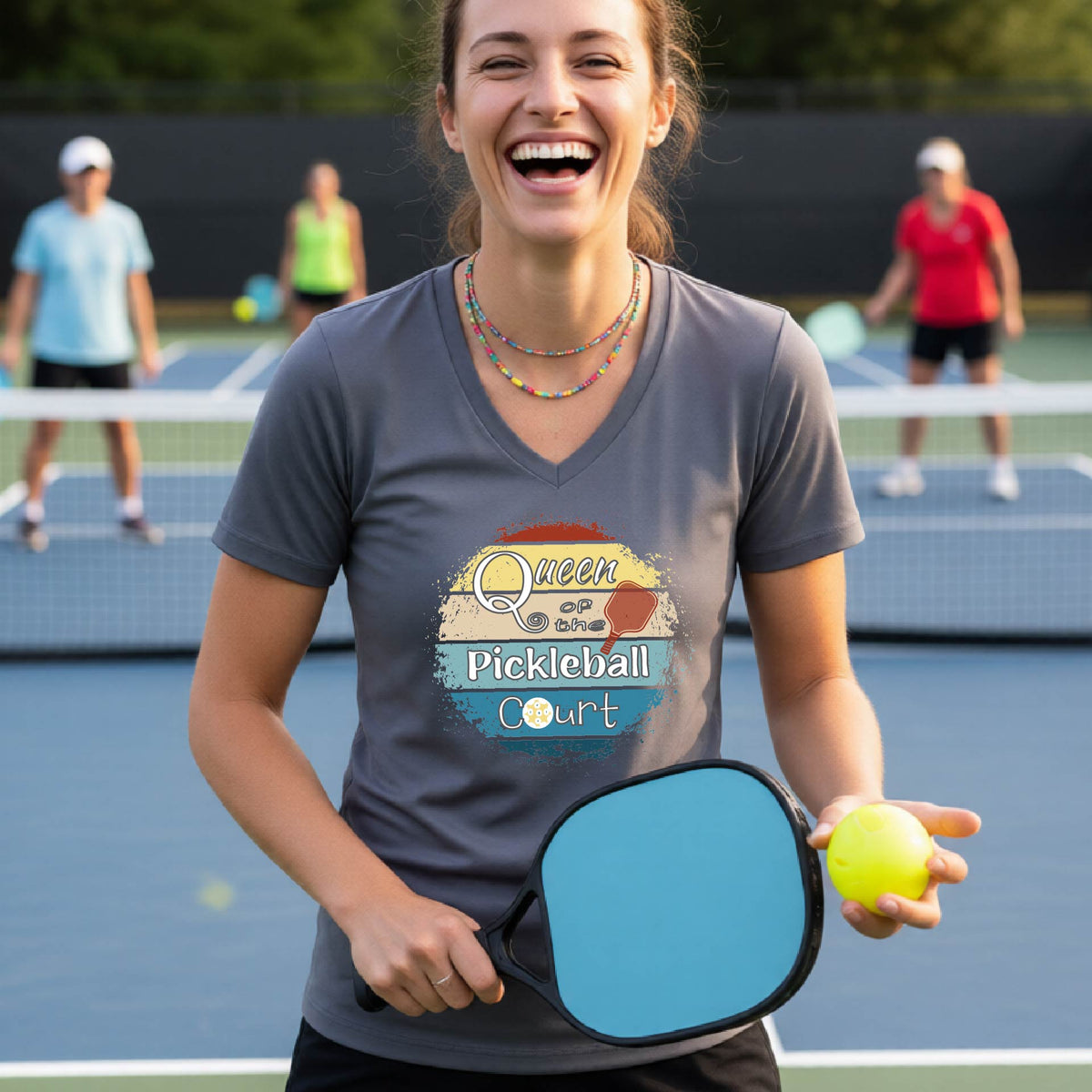 A woman on a pickleball court wearing a gray V-neck t-shirt with a colorful retro graphic that reads 'Queen Pickleball Court'. She is holding a blue paddle and a yellow pickleball, with other players blurred in the background.