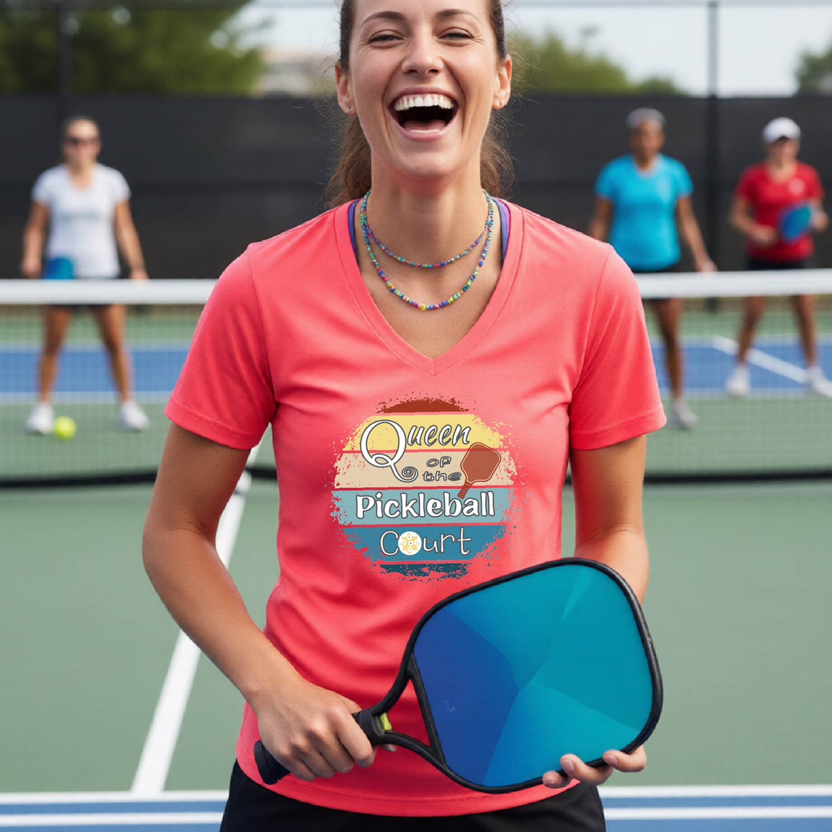 A woman on a pickleball court wearing a coral pink V-neck t-shirt with a 'Queen of the Pickleball Court' retro sunset graphic, holding a paddle and smiling. Other players are visible in the background on the court.