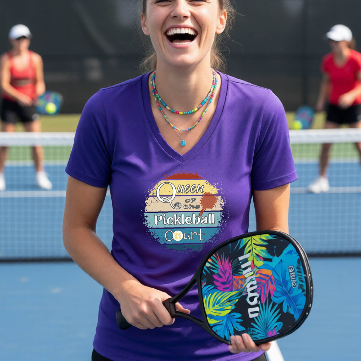 A woman on a pickleball court wearing a purple V-neck t-shirt with a 'Queen of the Pickleball Court' retro sunset graphic, holding a paddle and smiling. Other players are visible in the background on the court.