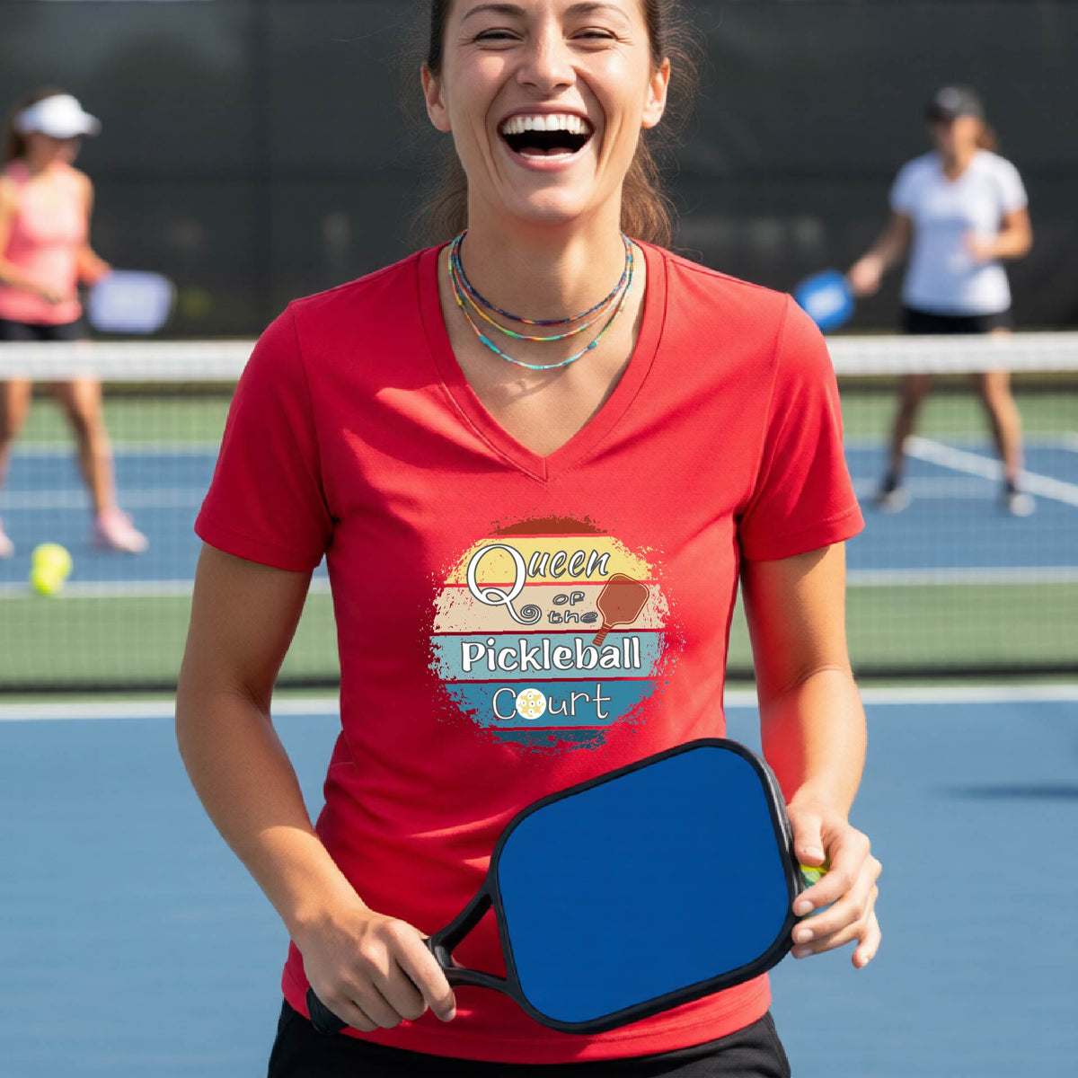 A woman on a pickleball court wearing a black V-neck t-shirt with a 'Queen of the Pickleball Court' retro sunset graphic, holding a paddle and smiling. Other players are visible in the background on the court.