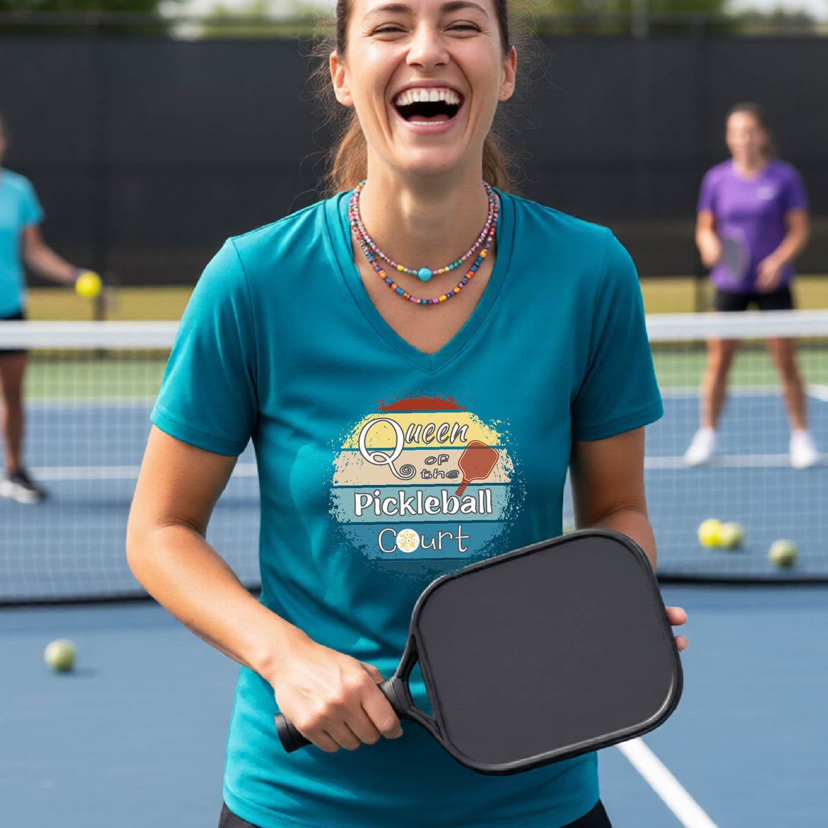 A woman laughing on a pickleball court wearing a teal graphic t-shirt that reads 'Queen Pickleball Court' with a retro pickleball design. She is holding a black pickleball paddle in the foreground, with other players and the court visible in the background.