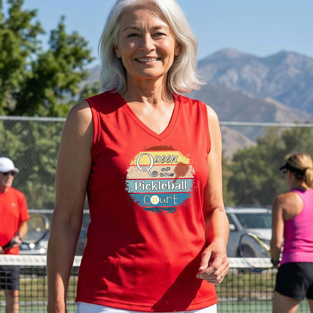 A woman on a pickleball court wearing a red sleeveless tank top with a colorful graphic that reads 'Queen of the Pickleball Court' in a retro sunset illustration.