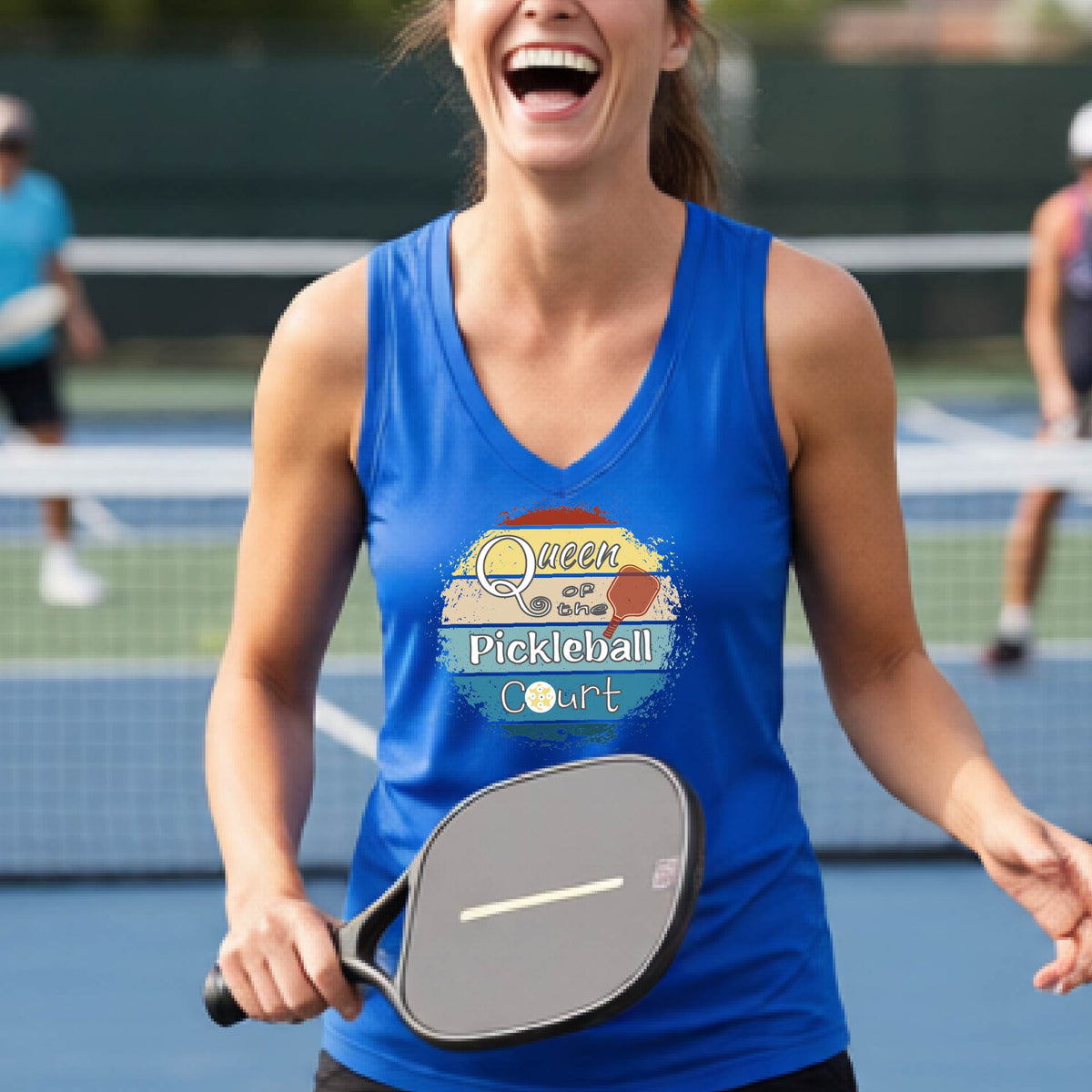 A woman on a pickleball court wearing a royal blue tank top with a colorful graphic that reads 'Queen of the Pickleball Court' in a retro sunset illustration.