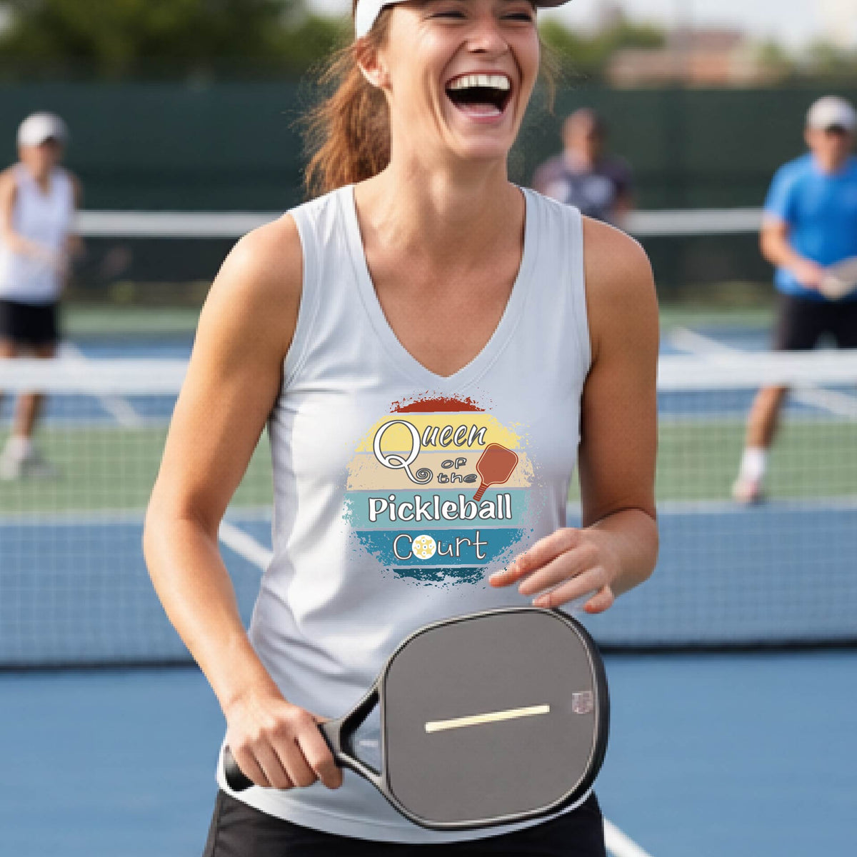 A woman on a pickleball court wearing a light silver gray tank top with a colorful graphic that reads 'Queen of the Pickleball Court' in a retro sunset illustration.