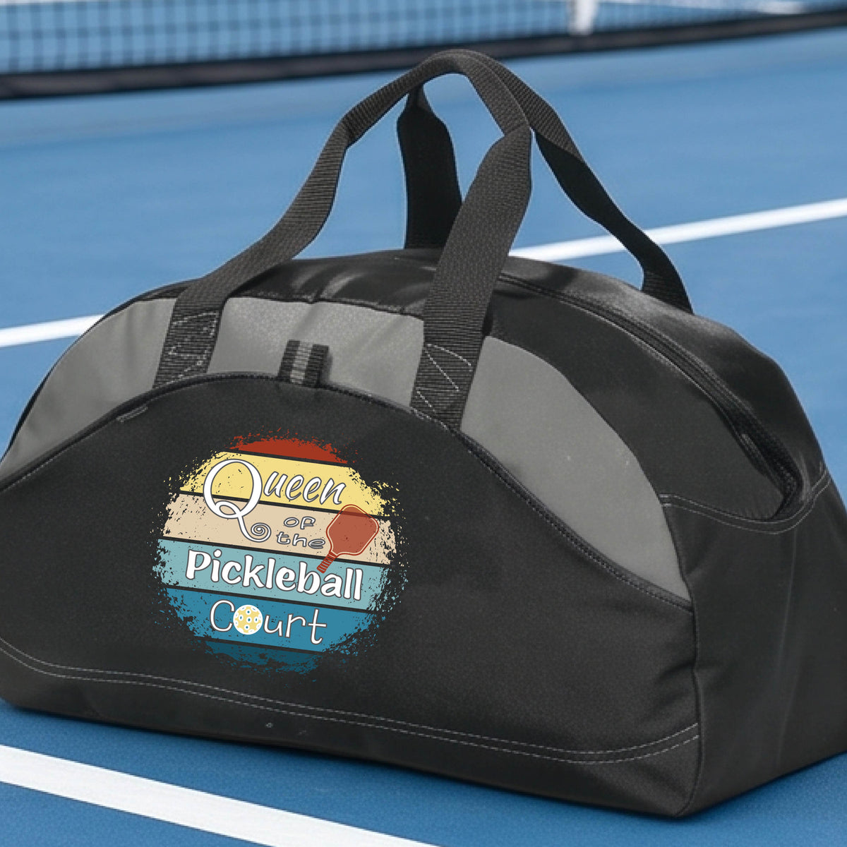 A custom pickleball duffel bag on a pickleball court, featuring a black color-blocked design with gray accents. The front has a custom graphic that reads 'Queen of hte Pickleball Court' with a pickleball illustration.