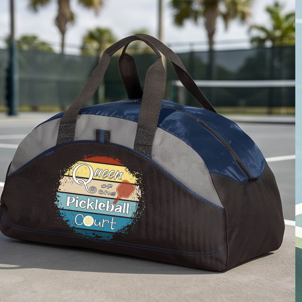 A custom pickleball duffel bag on a pickleball court, featuring a black and navy blue color-blocked design with gray accents. The front has a custom graphic that reads 'Queen of hte Pickleball Court' with a pickleball illustration.