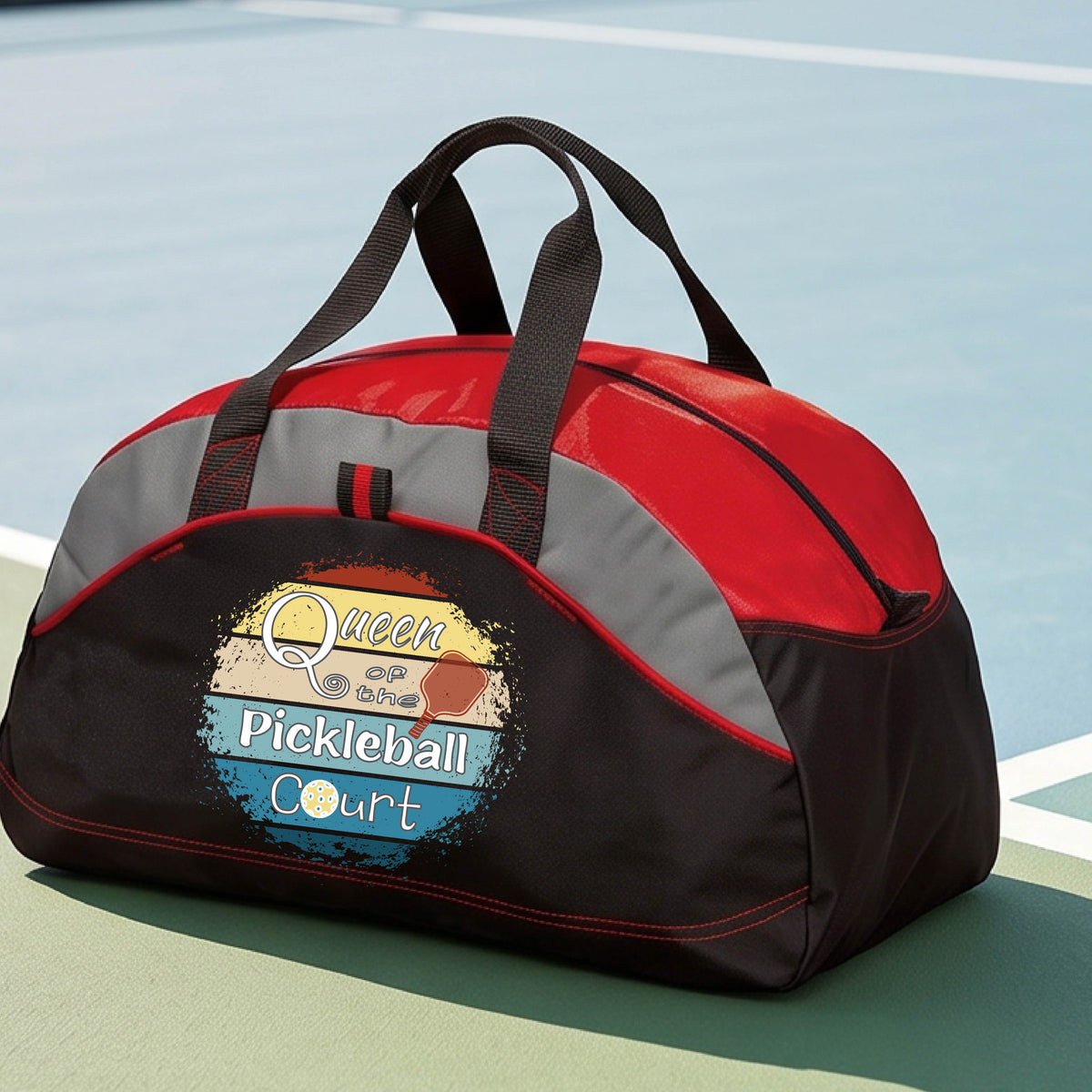 A custom pickleball duffel bag on a pickleball court, featuring a black and red color-blocked design with gray accents. The front has a custom graphic that reads 'Queen of hte Pickleball Court' with a pickleball illustration.