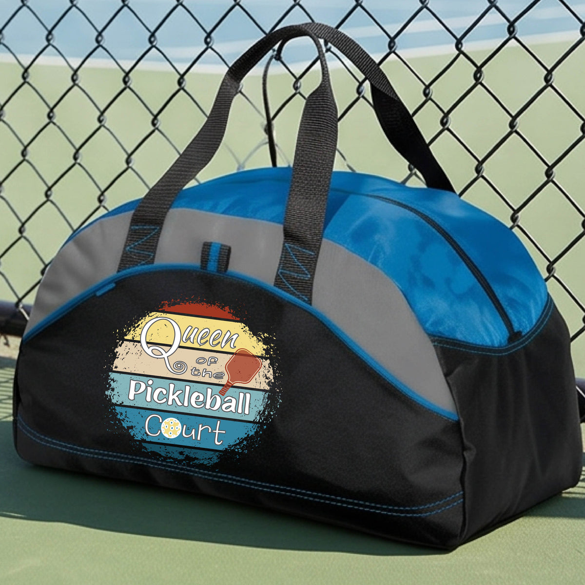 A custom pickleball duffel bag on a pickleball court, featuring a black and blue color-blocked design with gray accents. The front has a custom graphic that reads 'Queen of hte Pickleball Court' with a pickleball illustration.