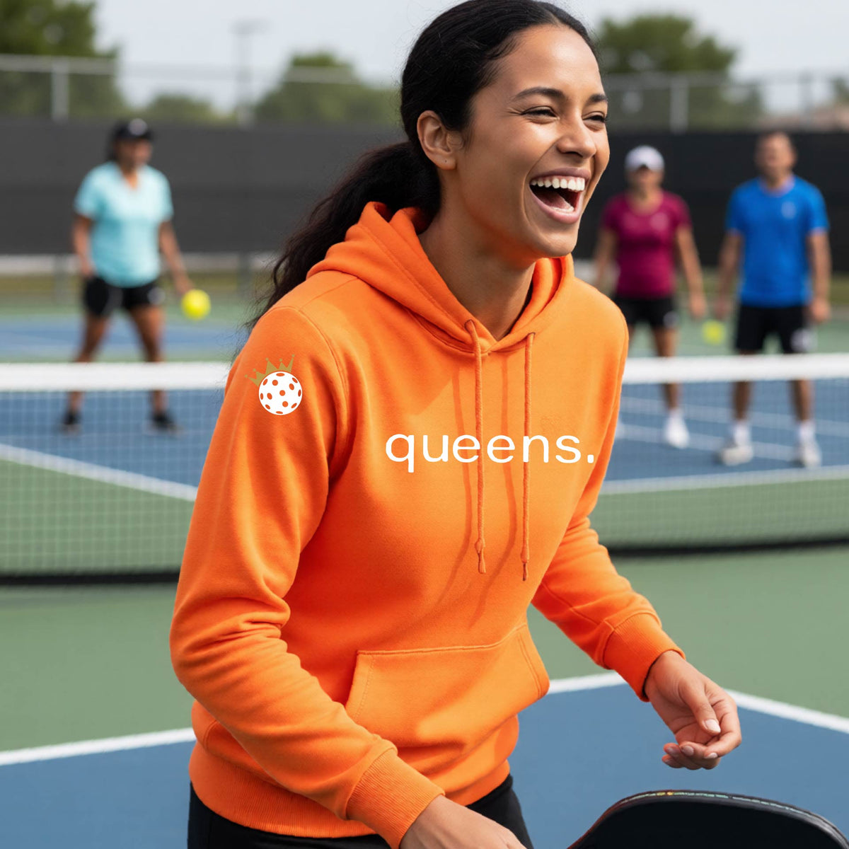 A woman laughing on an outdoor pickleball court wearing an orange hoodie with a white 'queens.' logo and a small pickleball wearing a gold crown graphic on the left sleeve.