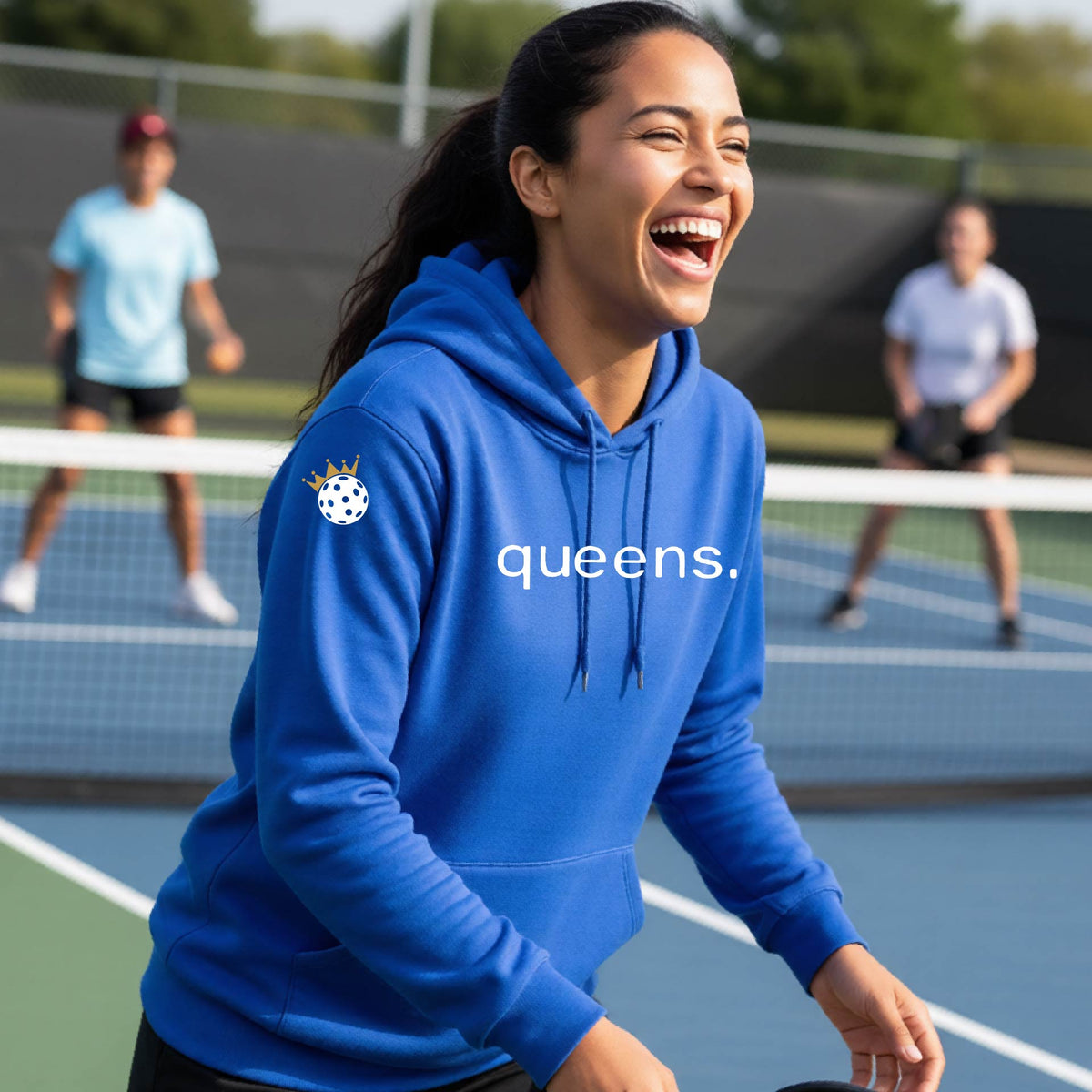 A woman laughing on an outdoor pickleball court wearing a royal blue hoodie with a white 'queens.' logo and a small pickleball wearing a gold crown graphic on the left sleeve.