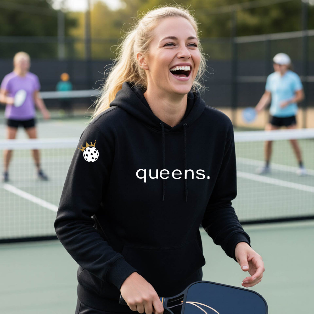 A woman laughing on an outdoor pickleball court wearing a black hoodie with a white 'queens.' logo and a small pickleball wearing a gold crown graphic on the left sleeve.