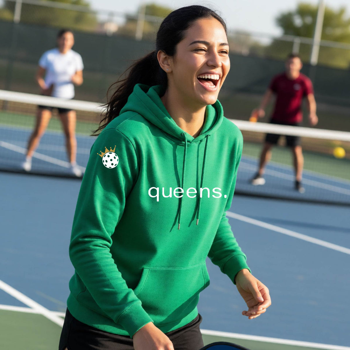 A woman laughing on an outdoor pickleball court wearing a green hoodie with a white 'queens.' logo and a small pickleball wearing a gold crown graphic on the left sleeve.