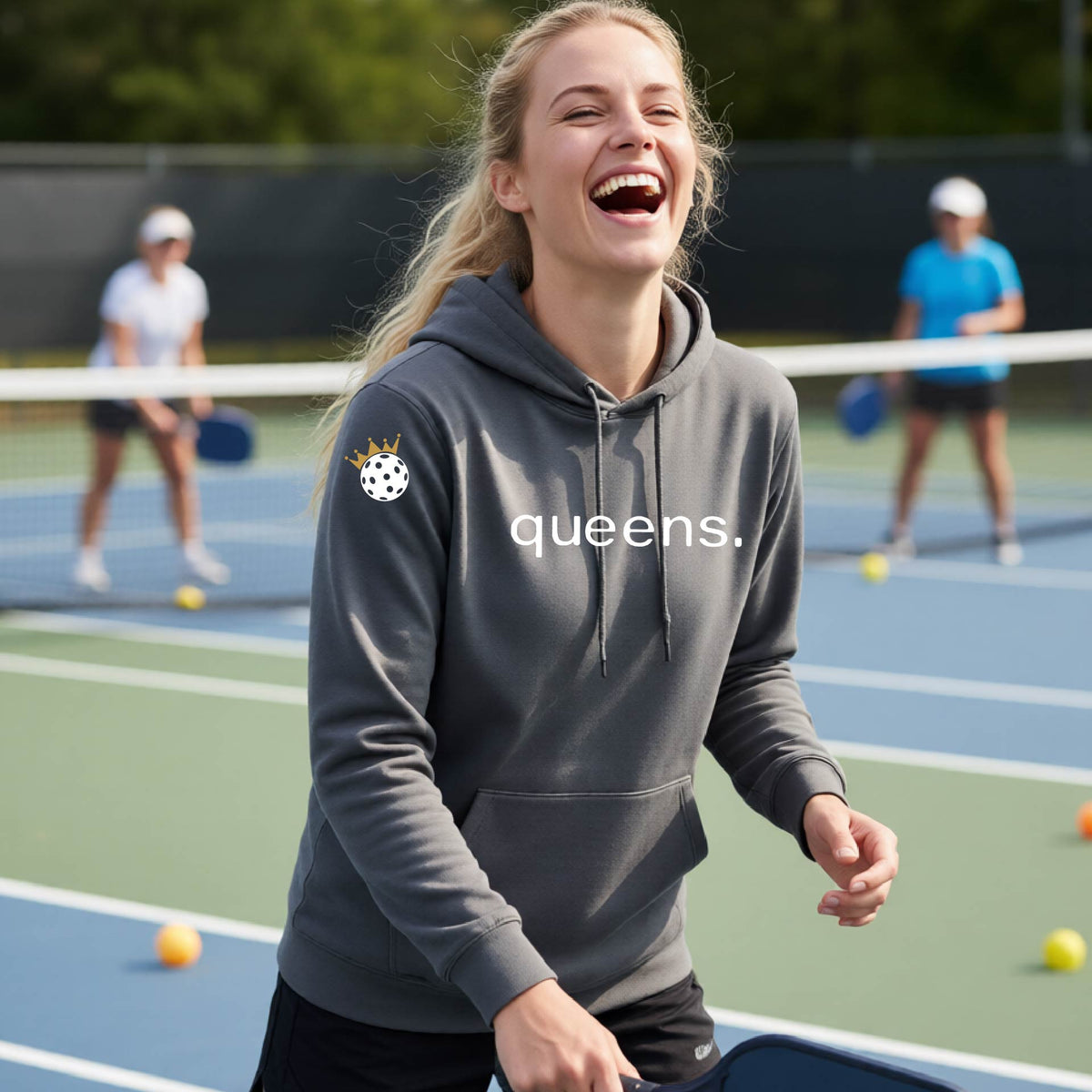 A woman laughing on an outdoor pickleball court wearing a gray hoodie with a white 'queens.' logo and a small pickleball wearing a gold crown graphic on the left sleeve.