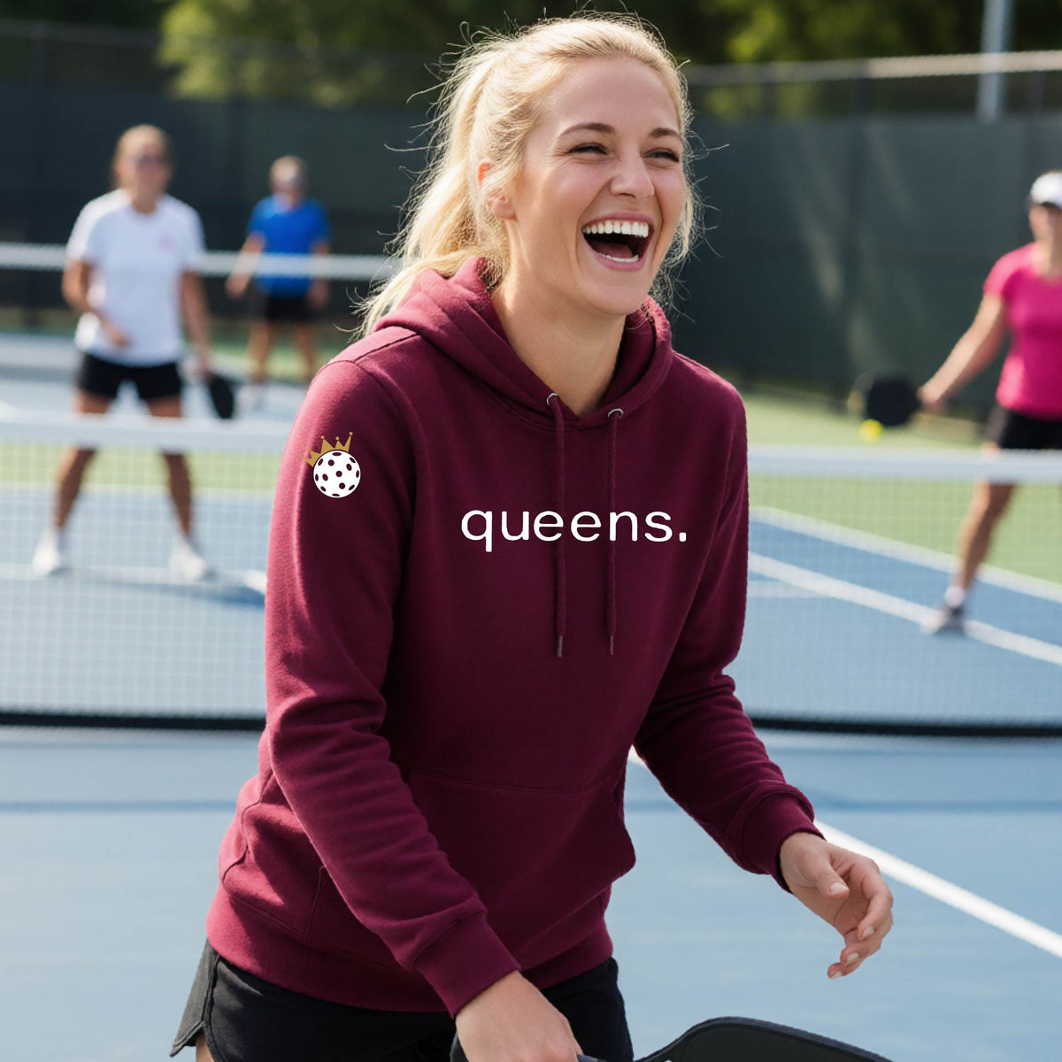A woman laughing on an outdoor pickleball court wearing a maroon hoodie with a white 'queens.' logo and a small pickleball wearing a gold crown graphic on the left sleeve.