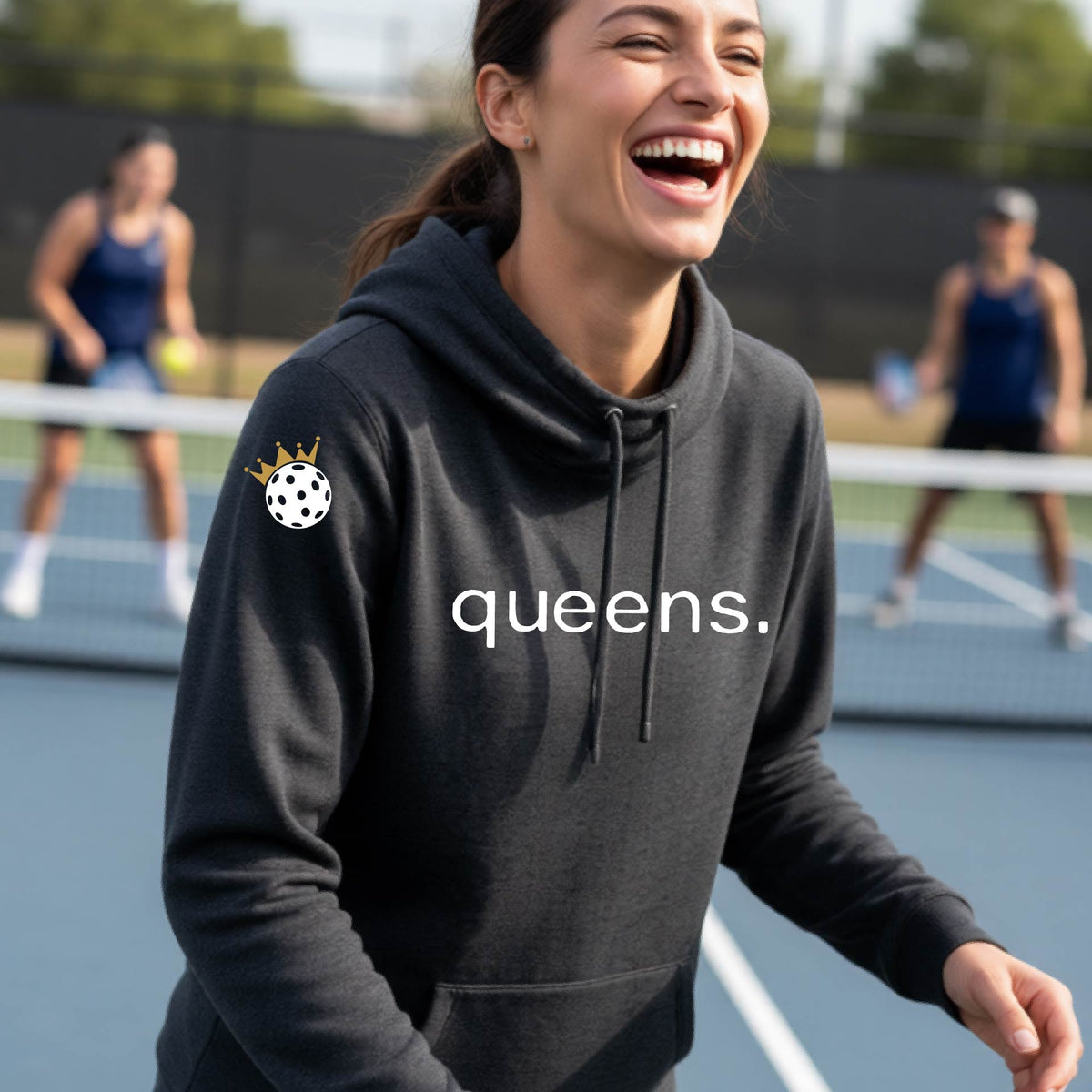 A woman laughing on a tennis court wearing a heather black cowl-neck hoodie with 'queens.' printed in white across the chest and a pickleball wearing a gold crown graphic on the sleeve.