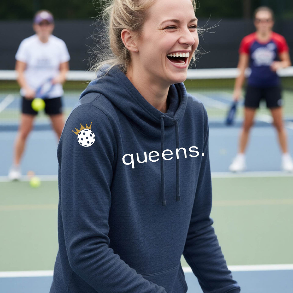 A woman laughing on a tennis court wearing a navy blue cowl-neck hoodie with 'queens.' printed in white across the chest and a pickleball wearing a gold crown graphic on the sleeve.