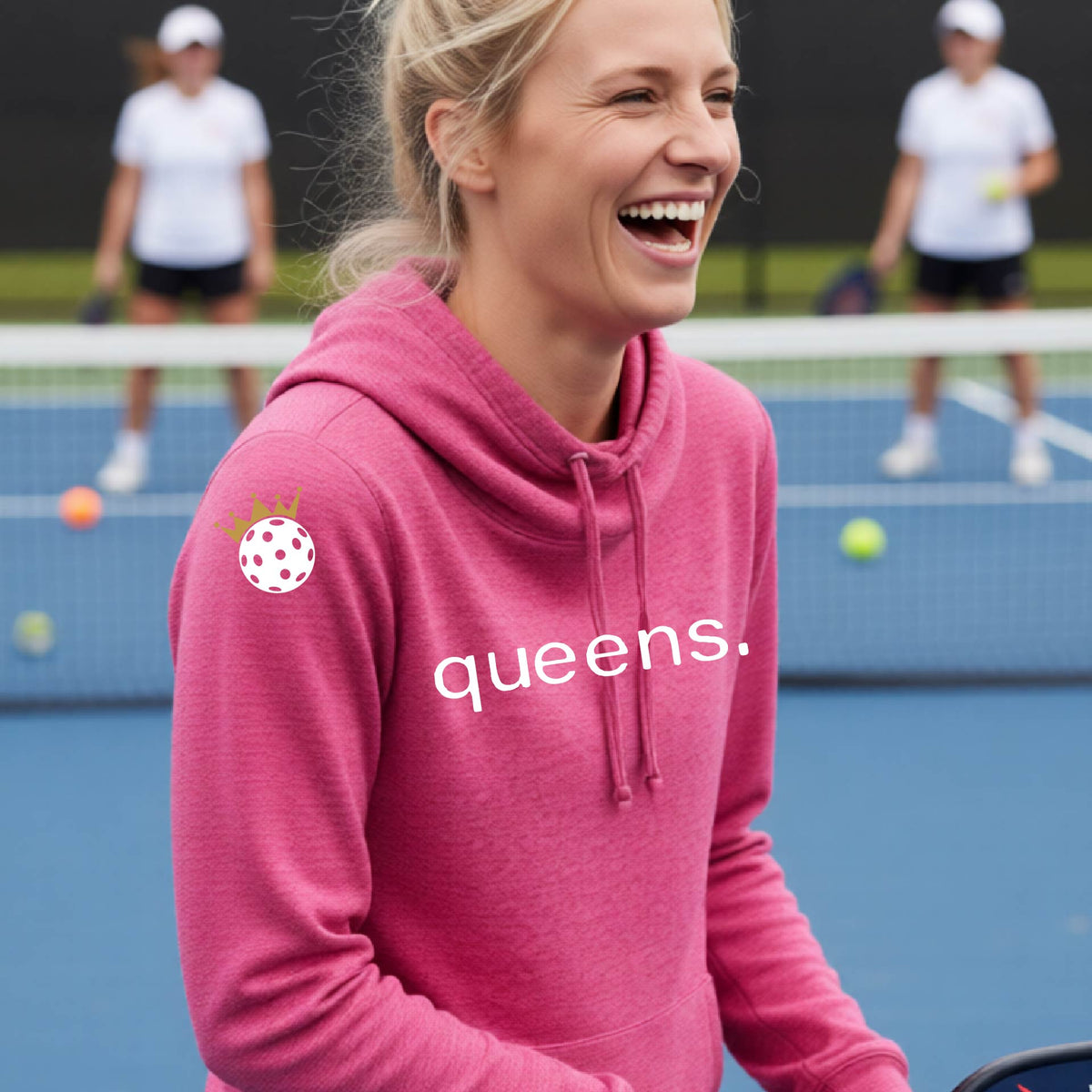 A woman laughing on a tennis court wearing a pink cowl-neck hoodie with 'queens.' printed in white across the chest and a pickleball wearing a gold crown graphic on the sleeve.