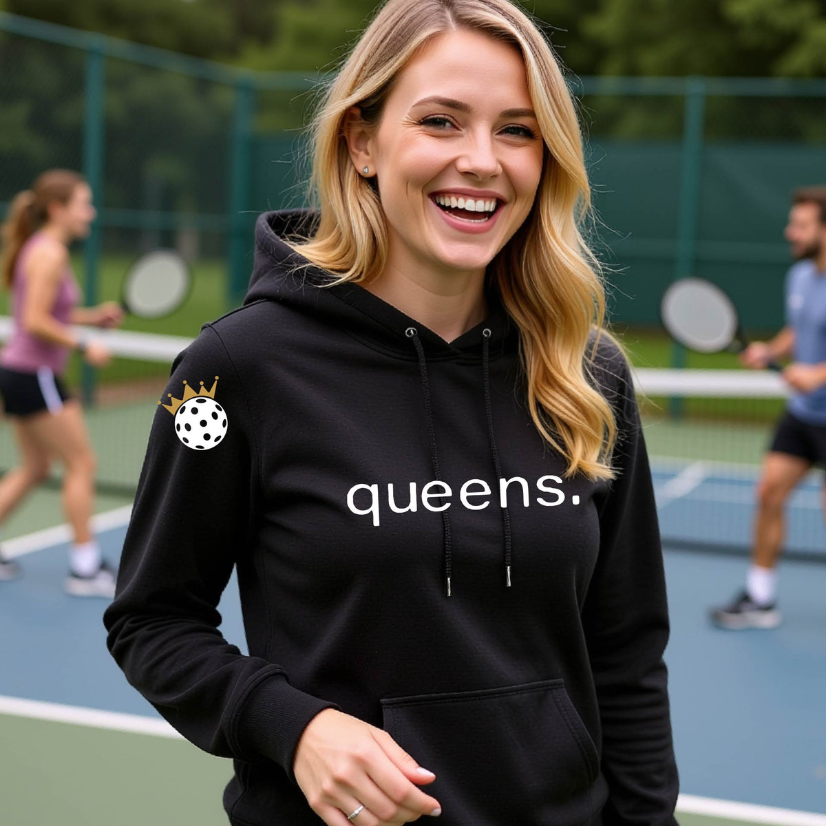 A smiling blonde woman wearing a black hoodie with 'queens.' printed in white across the chest and a small pickleball wearing a gold crown graphic on the sleeve, standing on an outdoor pickleball court. 