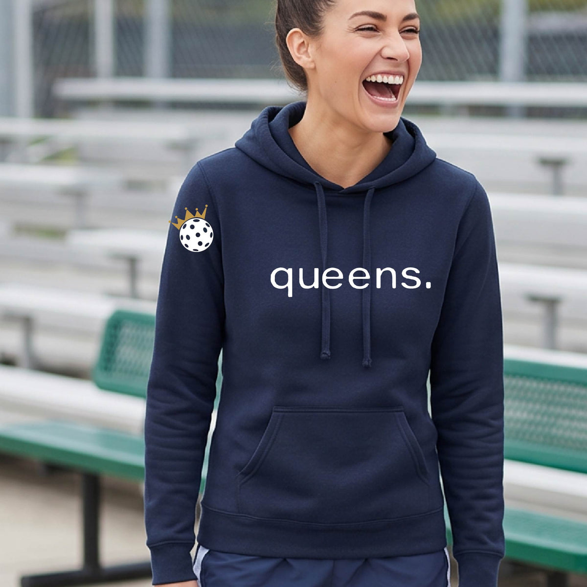 A smiling blonde woman wearing a navy blue hoodie with 'queens.' printed in white across the chest and a small pickleball wearing a gold crown graphic on the sleeve, standing on an outdoor pickleball court. 