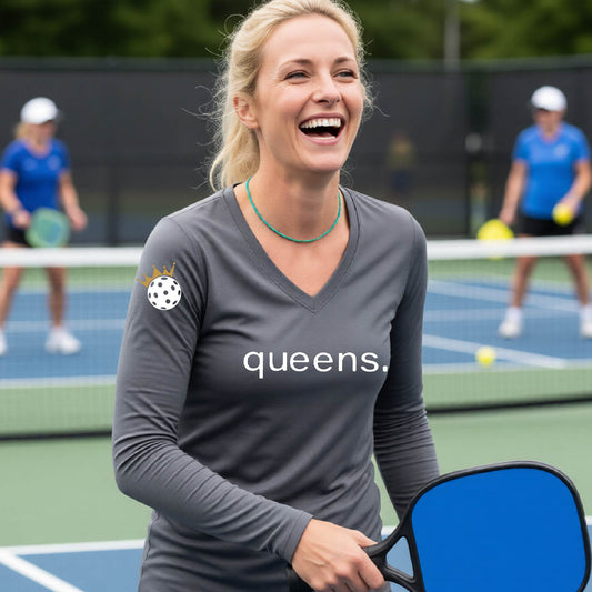 A woman laughing on a pickleball court wearing a dark gray long-sleeve V-neck shirt with 'queens,' printed in white across the chest and a small pickleball wearing a crown  on the sleeve.