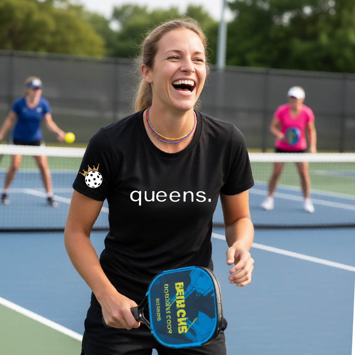 A woman laughing on a pickleball court wearing a bright black t-shirt with the word 'queens.' printed on the front, with a white pickleball wearing a crown on the left sleeve. 