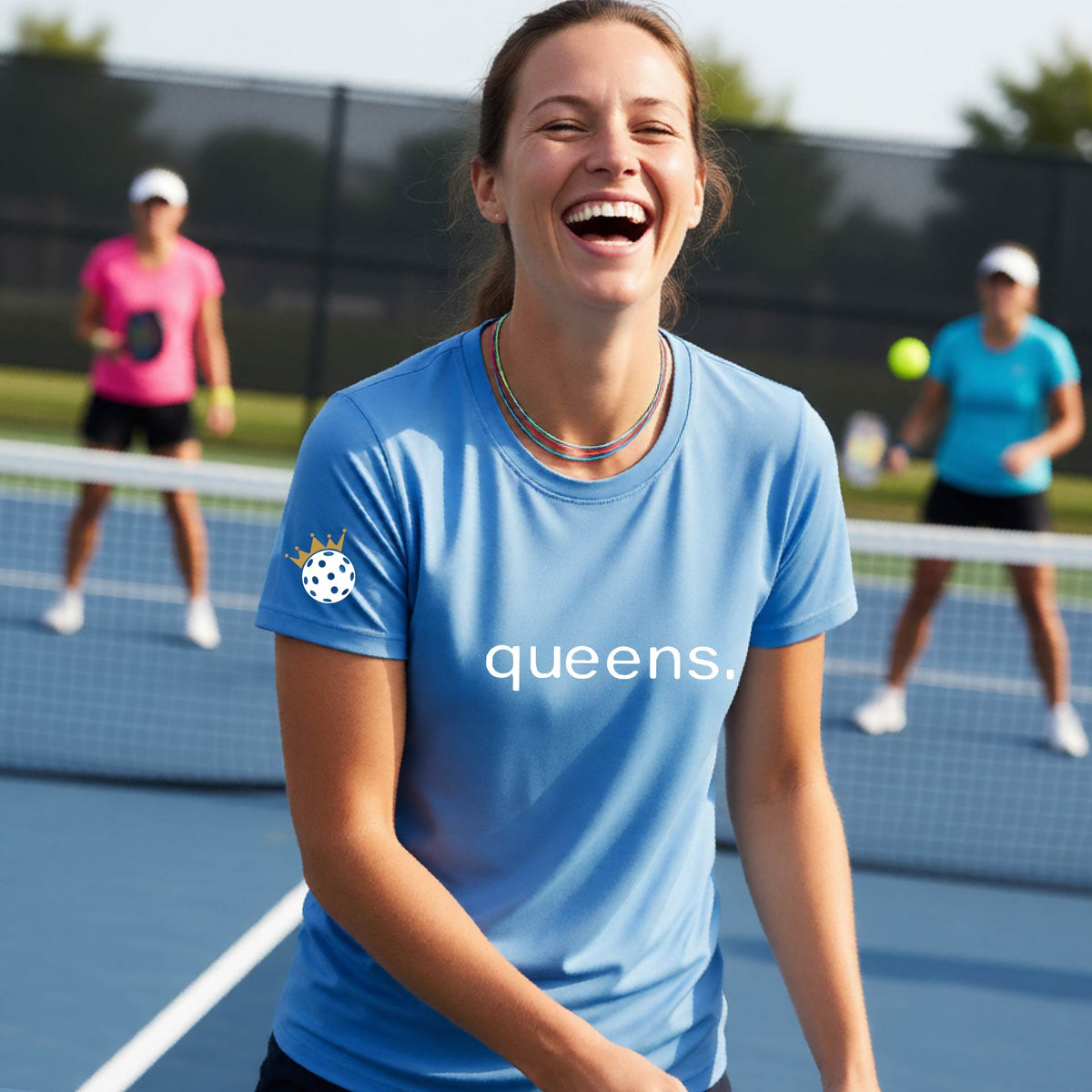 A woman laughing on a pickleball court wearing a bright blue t-shirt with the word 'queens.' printed on the front, with a white pickleball wearing a crown on the left sleeve. 