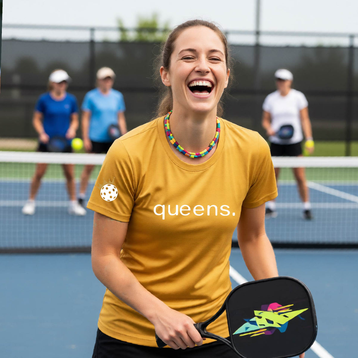 A woman laughing on a pickleball court wearing a bright mustard yellow gold  t-shirt with the word 'queens.' printed on the front, with a white pickleball wearing a crown on the left sleeve. 