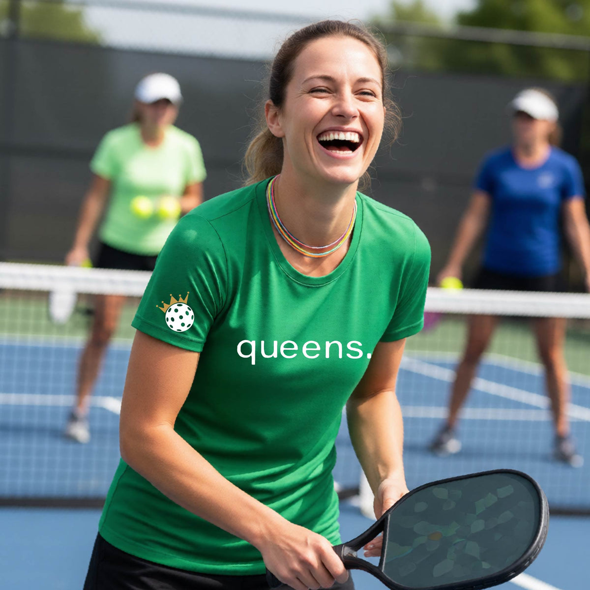 A woman laughing on a pickleball court wearing a green t-shirt with the word 'queens.' printed on the front, with a white pickleball wearing a crown on the left sleeve. 