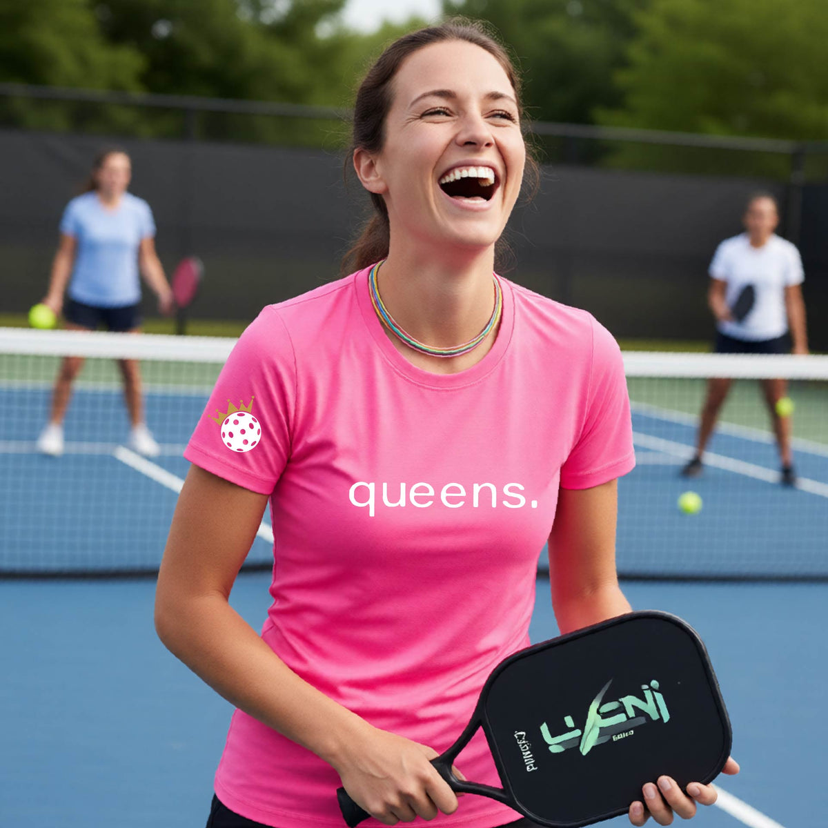 A woman laughing on a pickleball court wearing a bright pink t-shirt with the word 'queens.' printed on the front, with a white pickleball wearing a crown on the left sleeve. 