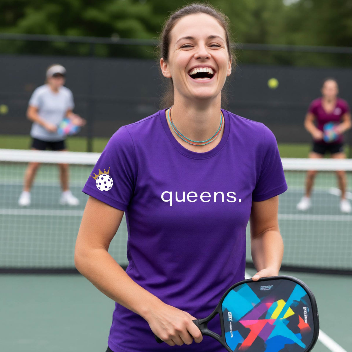 A woman laughing on a pickleball court wearing a bright purple t-shirt with the word 'queens.' printed on the front, with a white pickleball wearing a crown on the left sleeve. 