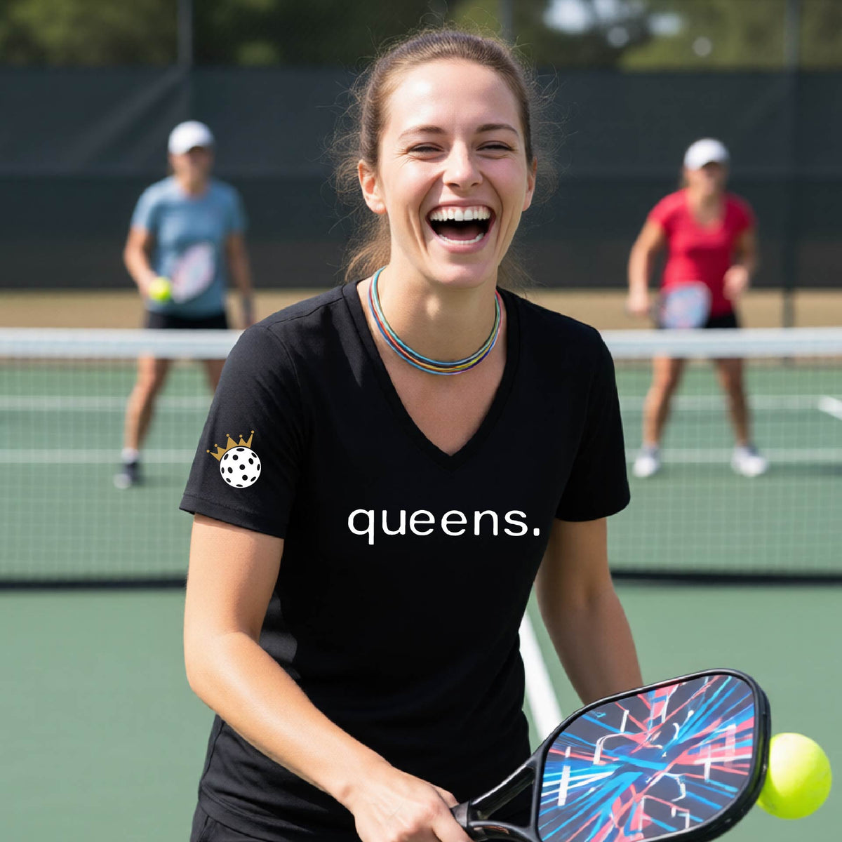 A woman laughing on a pickleball court wearing a short-sleeve black V-neck t-shirt with the word 'queens.' printed in white across the chest and a small lwhite pickleball wearing a gold crown on the sleeve.