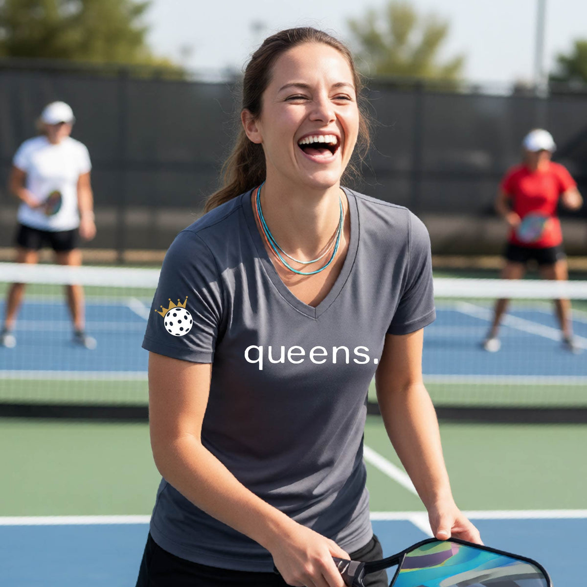 A woman laughing on a pickleball court wearing a short-sleeve dark gray  V-neck t-shirt with the word 'queens.' printed in white across the chest and a small lwhite pickleball wearing a gold crown on the sleeve.