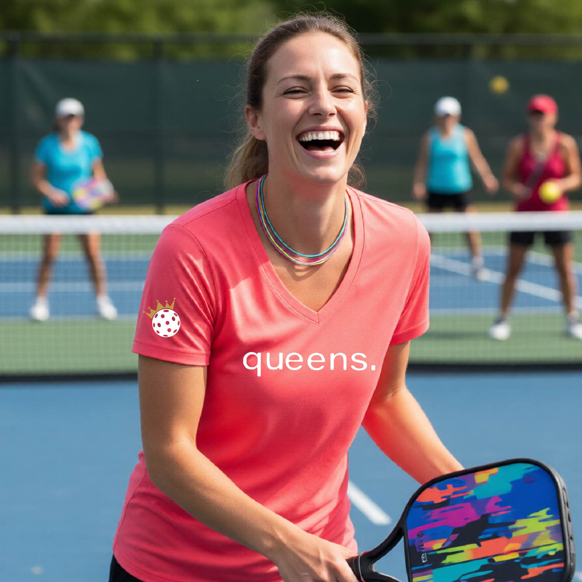 A woman laughing on a pickleball court wearing a short-sleeve coral pink V-neck t-shirt with the word 'queens.' printed in white across the chest and a small lwhite pickleball wearing a gold crown on the sleeve.
