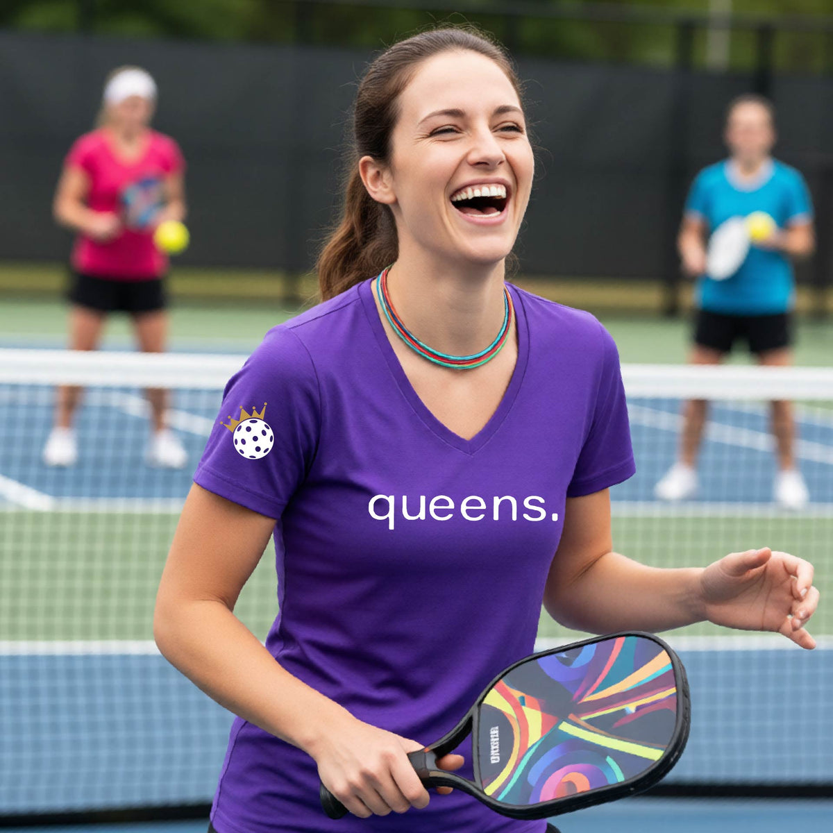 A woman laughing on a pickleball court wearing a short-sleeve purple V-neck t-shirt with the word 'queens.' printed in white across the chest and a small lwhite pickleball wearing a gold crown on the sleeve.