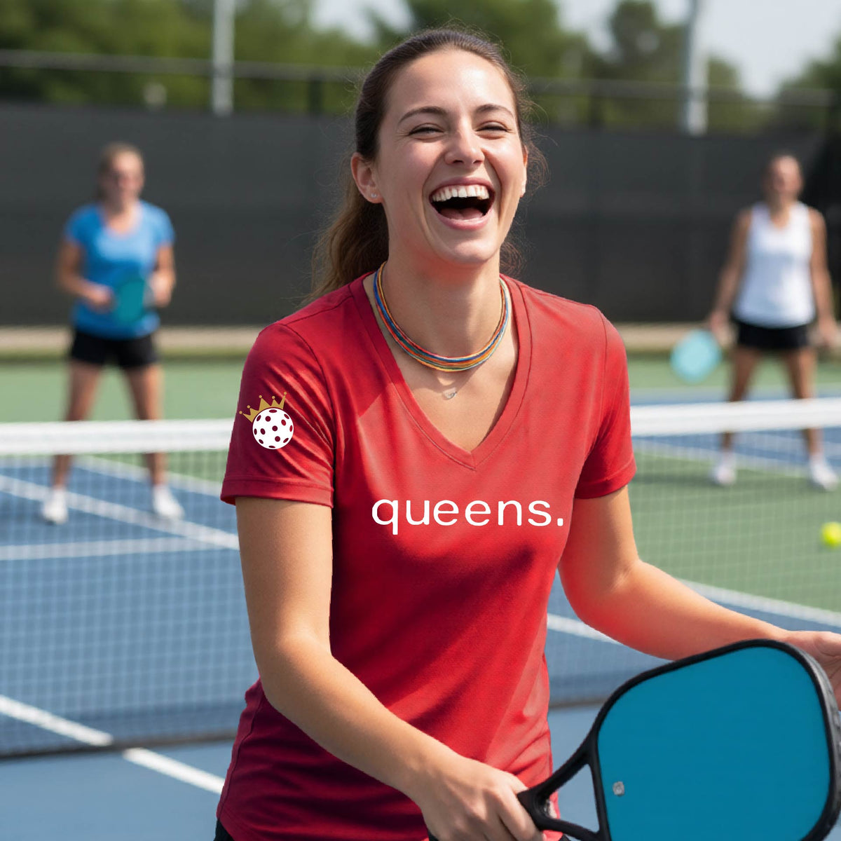 A woman laughing on a pickleball court wearing a short-sleeve red V-neck t-shirt with the word 'queens.' printed in white across the chest and a small lwhite pickleball wearing a gold crown on the sleeve.