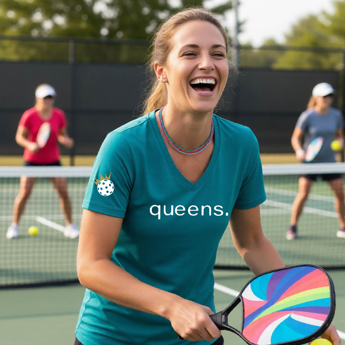A woman laughing on a pickleball court wearing a short-sleeve teal V-neck t-shirt with the word 'queens.' printed in white across the chest and a small lwhite pickleball wearing a gold crown on the sleeve.