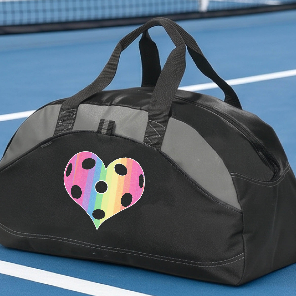 A close up of a medium size black duffel bag, with gray panels,  on a pickleball court, with a graphic of a brightly rainbow-colored heart designed like a pickleball on the front.