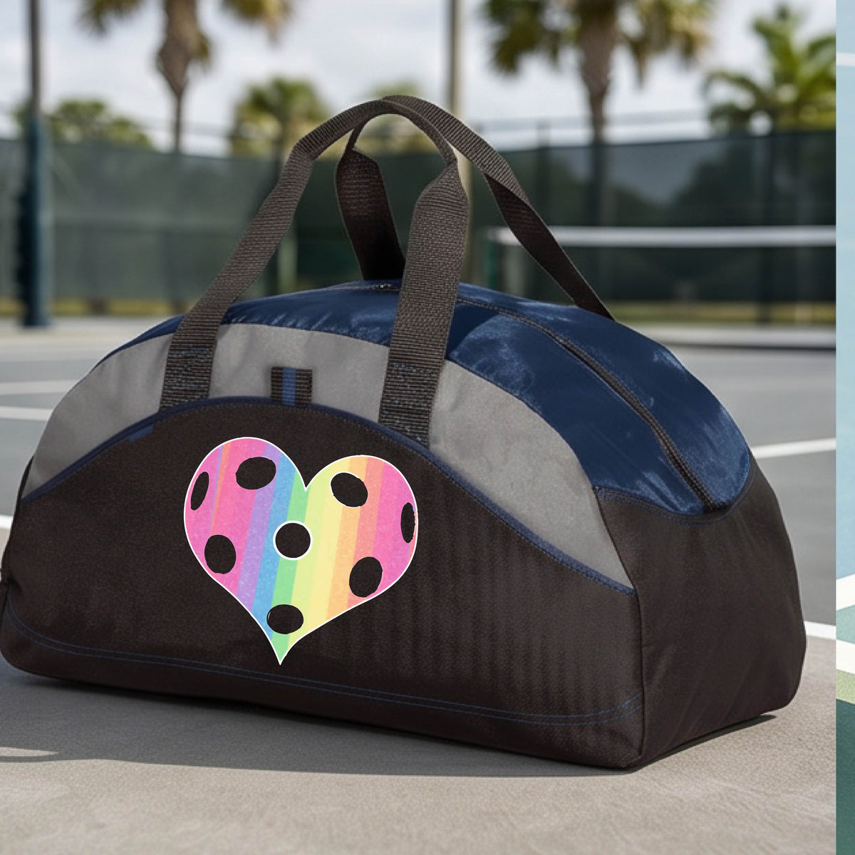 A close up of a medium size navy blue and black duffel bag, with gray panels,  on a pickleball court, with a graphic of a brightly rainbow-colored heart designed like a pickleball on the front.