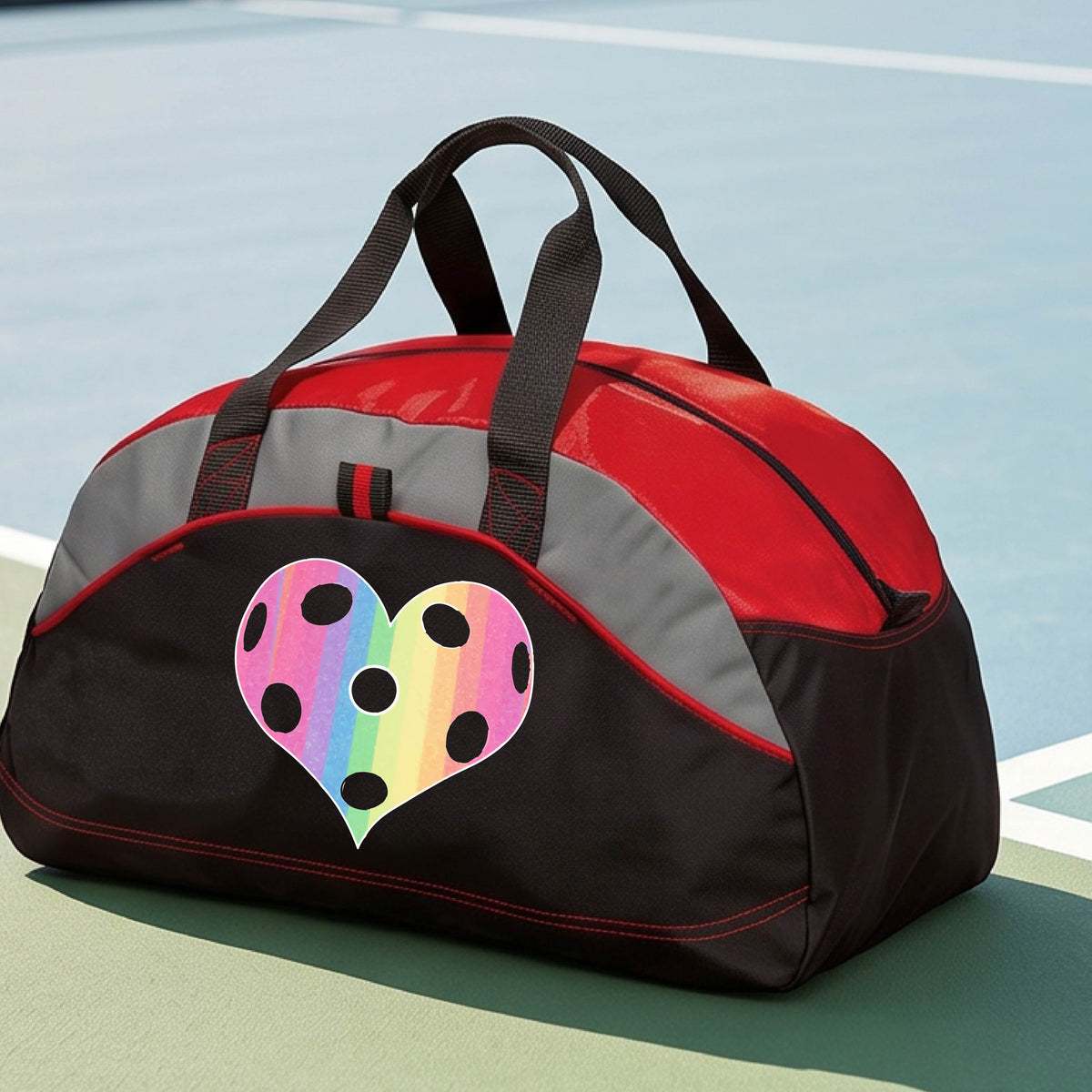 A close up of a medium size red and black duffel bag, with gray panels,  on a pickleball court, with a graphic of a brightly rainbow-colored heart designed like a pickleball on the front.