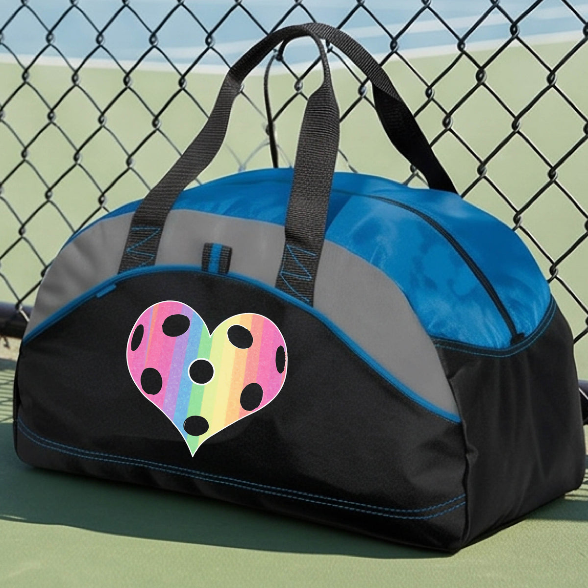 A close up of a medium size royal blue and black duffel bag, with gray panels,  on a pickleball court, with a graphic of a brightly rainbow-colored heart designed like a pickleball on the front.