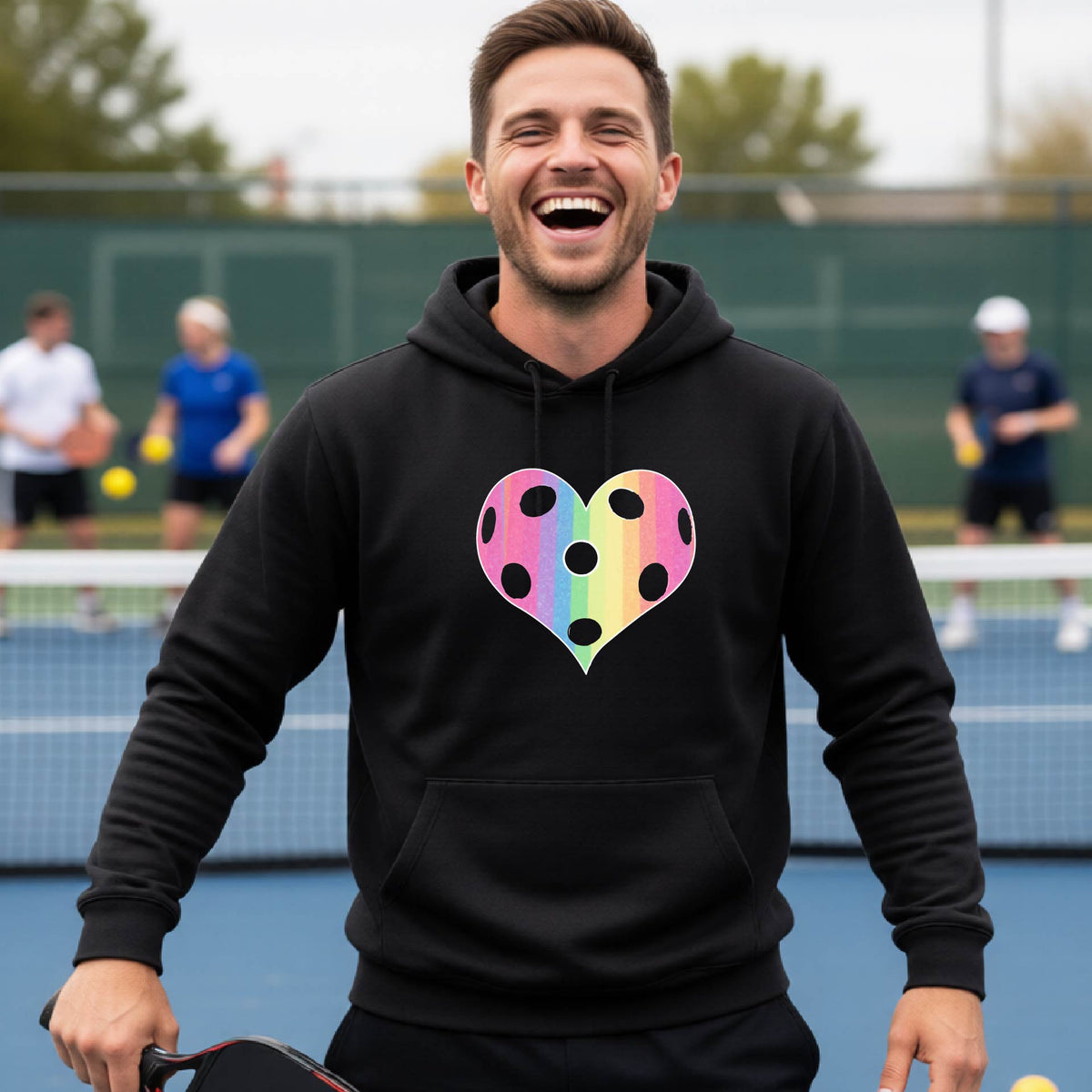 A man laughing on a pickleball court, wearing a black unisex hoodie pullover with a graphic of a brightly rainbow-colored heart designed like a pickleball.