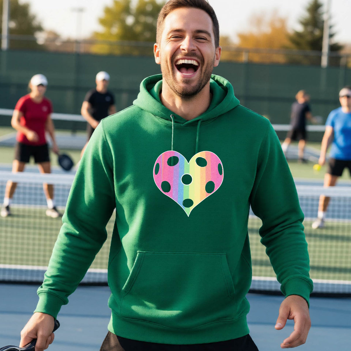 A man laughing on a pickleball court, wearing a green unisex hoodie pullover with a graphic of a brightly rainbow-colored heart designed like a pickleball.