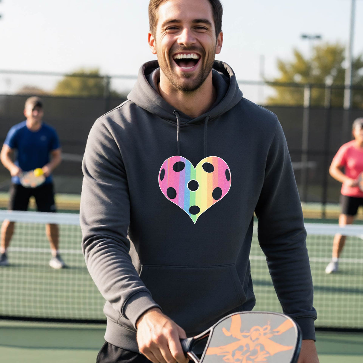 A man laughing on a pickleball court, wearing a dark gray unisex hoodie pullover with a graphic of a brightly rainbow-colored heart designed like a pickleball.