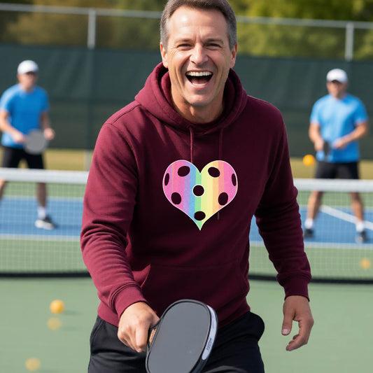 A man laughing on a pickleball court, wearing a maroon unisex hoodie pullover with a graphic of a brightly rainbow-colored heart designed like a pickleball.