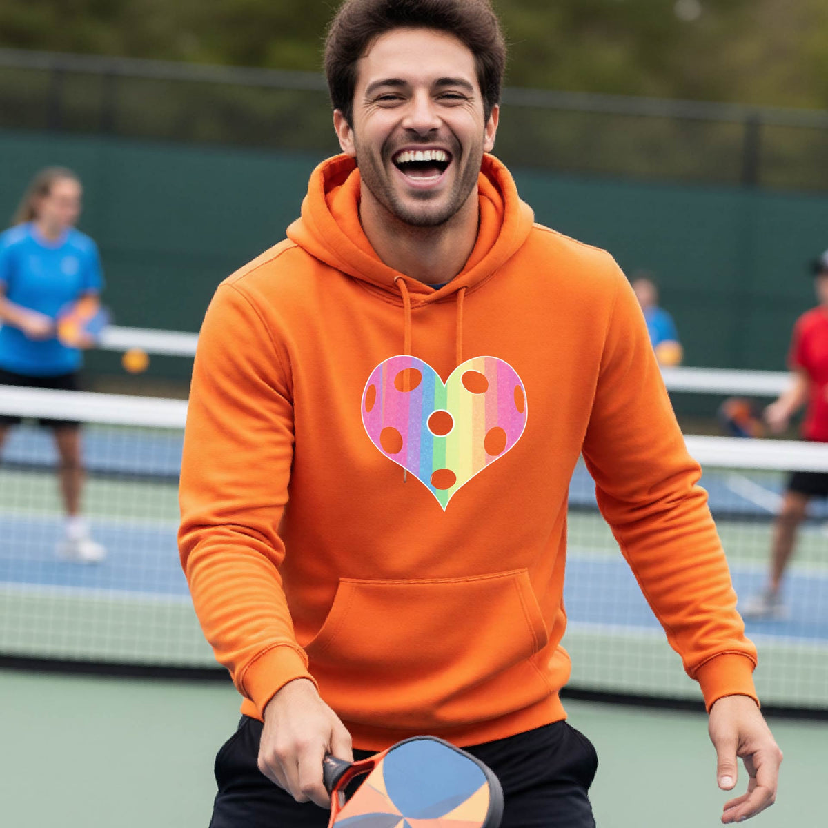A man laughing on a pickleball court, wearing an orange unisex hoodie pullover with a graphic of a brightly rainbow-colored heart designed like a pickleball.