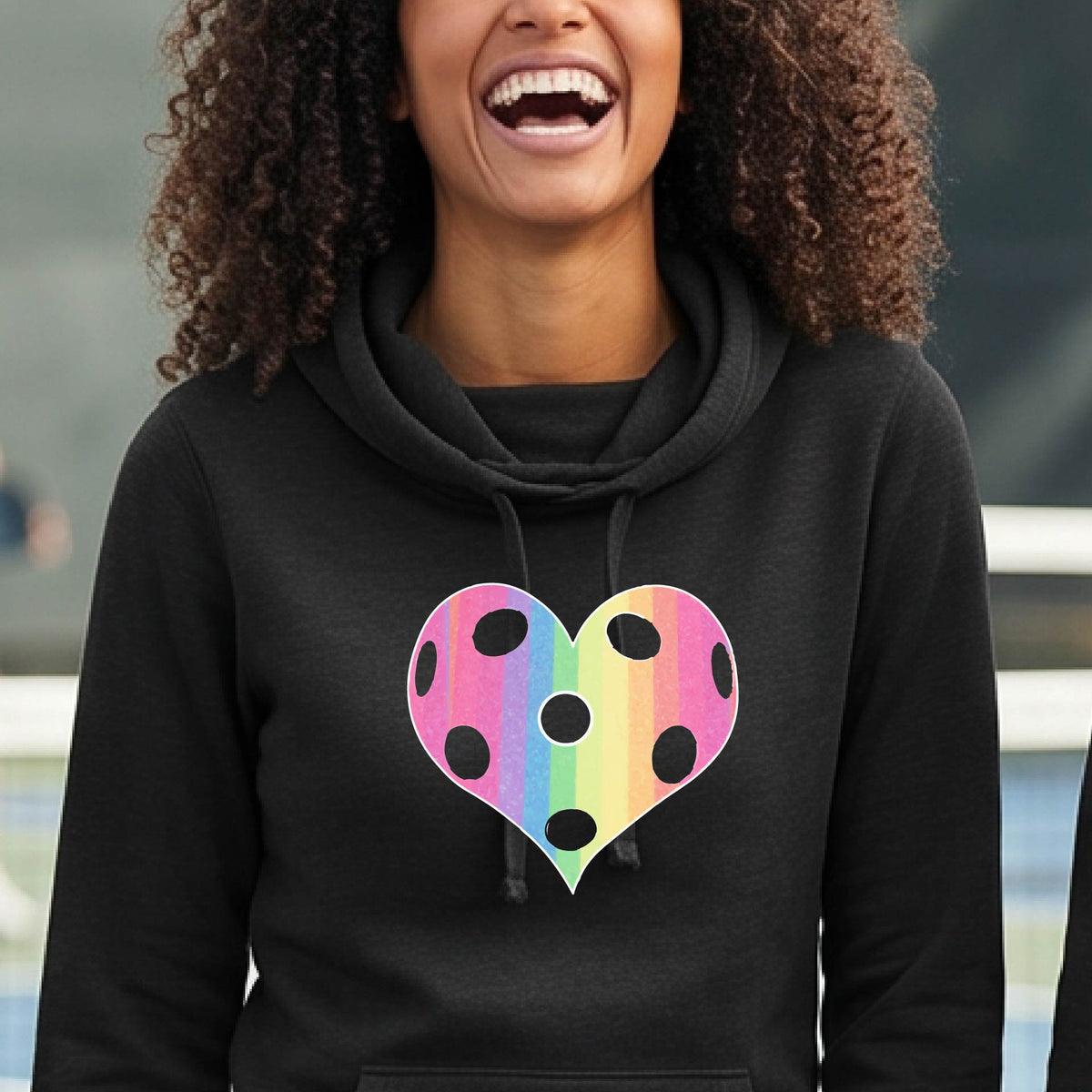 A woman laughing on a pickleball court, wearing a black cowl-neck hoodie pullover with a graphic of a brightly rainbow-colored heart designed like a pickleball.