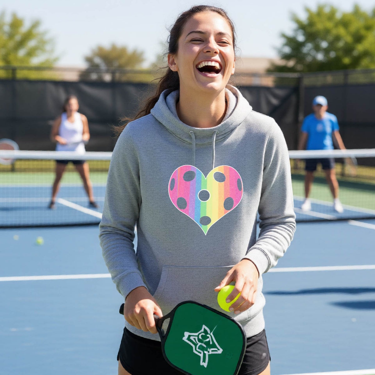 A woman laughing on a pickleball court, wearing a light gray cowl-neck hoodie pullover with a graphic of a brightly rainbow-colored heart designed like a pickleball.