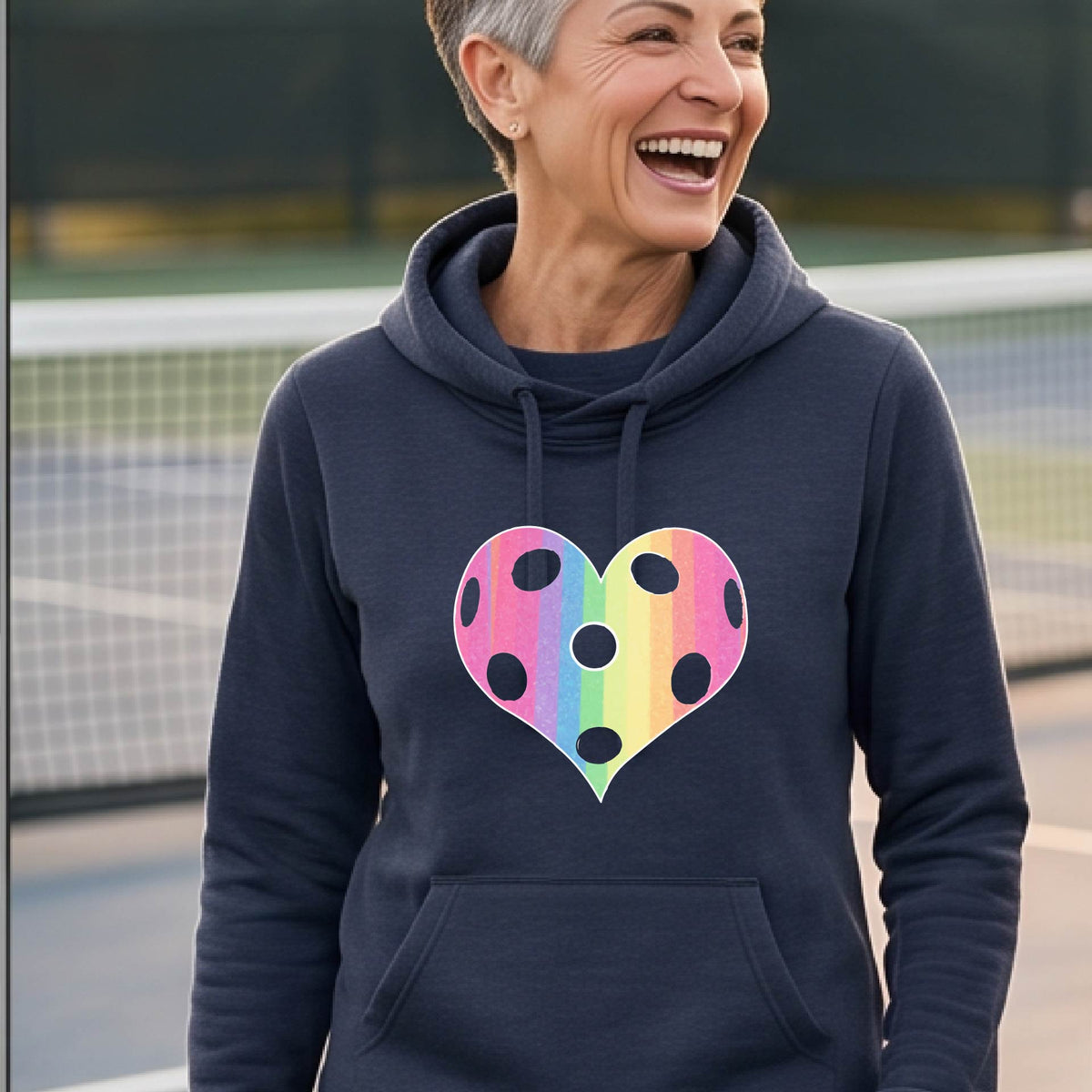 A woman laughing on a pickleball court, wearing a navy blue cowl-neck hoodie pullover with a graphic of a brightly rainbow-colored heart designed like a pickleball.